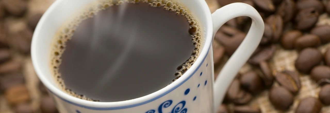 Taza de café caliente con vapor, rodeada de granos de café tostados sobre una superficie de yute.