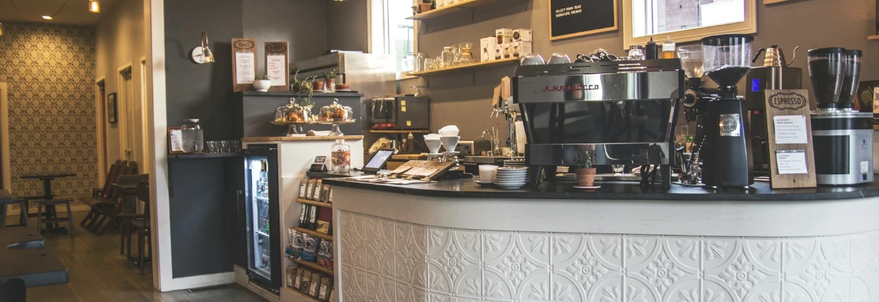 Barra de café moderna en una cafetería, con una máquina de espresso brillante, estantes con utensilios y una vitrina con productos. El ambiente es acogedor y contemporáneo, con decoración de fondo en tonos neutros y detalles decorativos en las paredes.