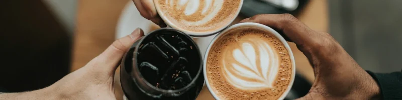 Tres tazas de café en manos de diferentes personas, con diseños de latte art en dos tazas y una bebida fría en la tercera, sobre una mesa de madera.
